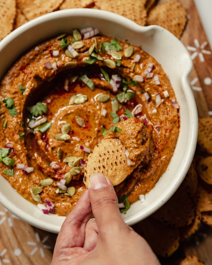 An overhead shot of a hand swiping a tortilla chip through a deep red dip that's garnished with cilantro, olive oil, and finely diced red onion.