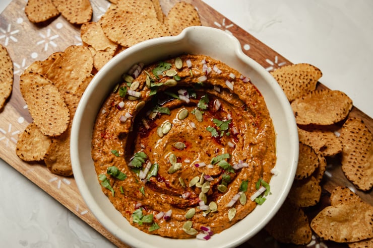 An overhead shot of a deep red black bean dip that is garnished with pumpkin seeds, finely diced red onion, olive oil, and cilantro. Tortilla chips are seen to the side.