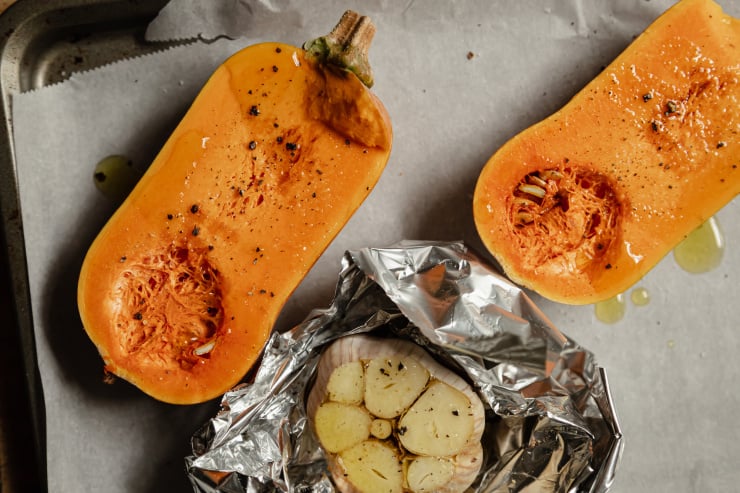 An overhead shot of honeynut squash halves and a bulb of garlic on a baking sheet. All is drizzled with olive oil.