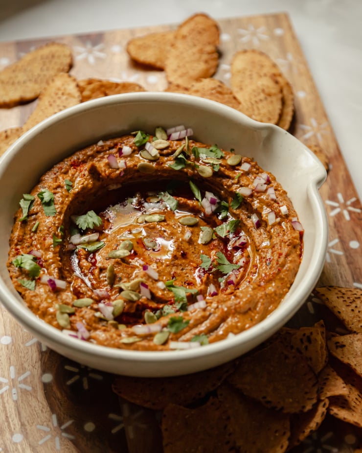 A 3/4 angle shot of a deep red dip in an off white bowl. The dip is garnished with pumpkin seeds, cilantro, olive oil, and red onion.