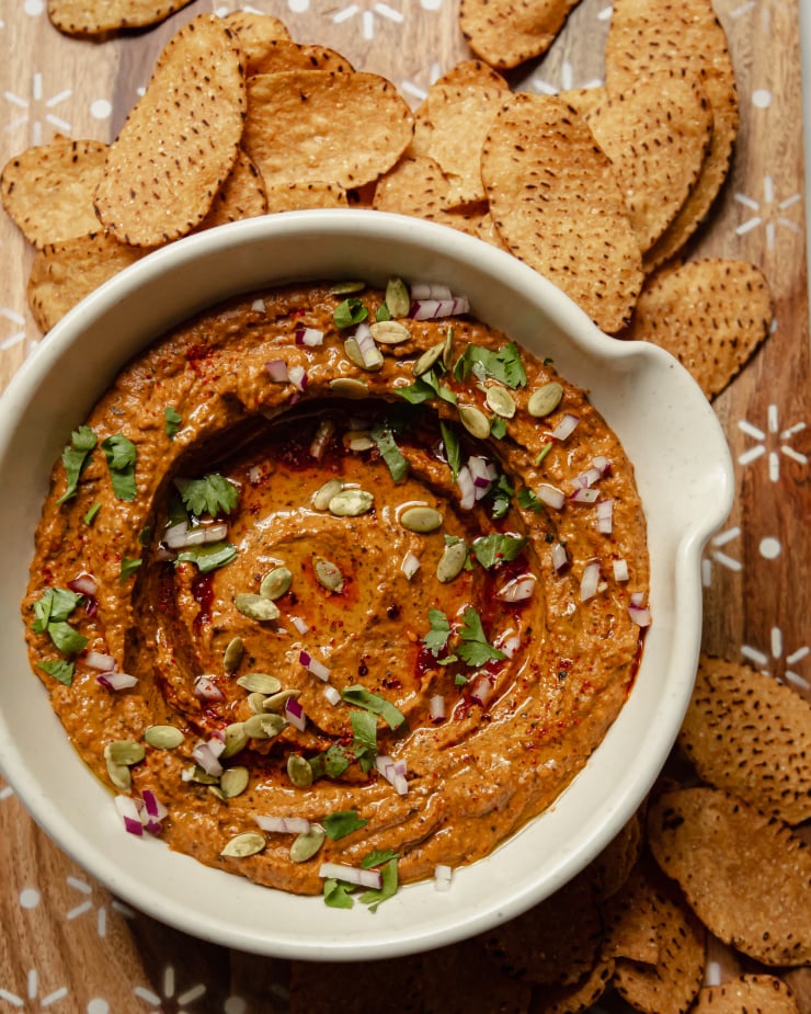 An overhead shot of a deep red black bean dip that is garnished with pumpkin seeds, finely diced red onion, olive oil, and cilantro. Tortilla chips are seen to the side.