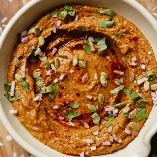 An up close, overhead shot of a deep red black bean dip that is garnished with pumpkin seeds, finely diced red onion, olive oil, and cilantro.