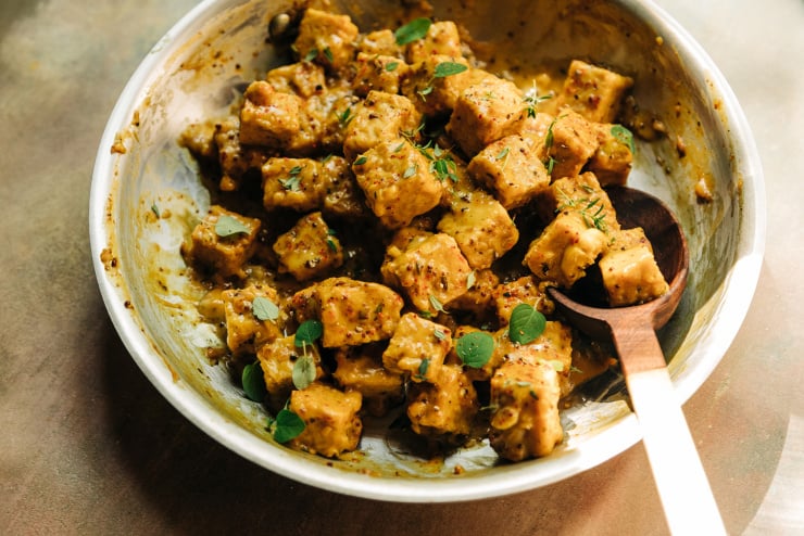 A 3/4 angle shot of saucy chunks of maple mustard tempeh in a skillet. The sauce is golden yellow and the tempeh is garnished with fresh thyme and oregano leaves.
