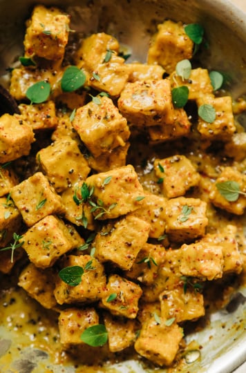 An up close, overhead shot of saucy chunks of tempeh in a skillet. The sauce is golden yellow and the tempeh is garnished with fresh thyme and oregano leaves.