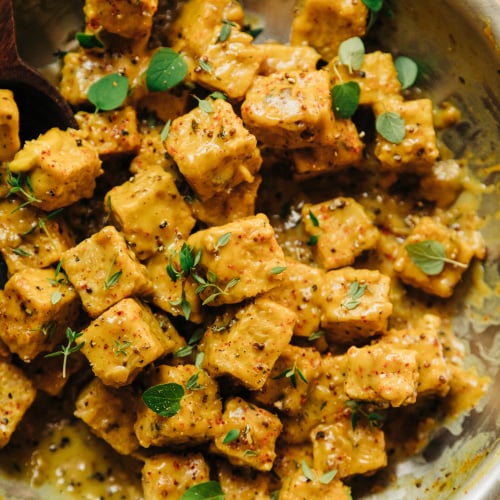 An up close, overhead shot of saucy chunks of tempeh in a skillet. The sauce is golden yellow and the tempeh is garnished with fresh thyme and oregano leaves.