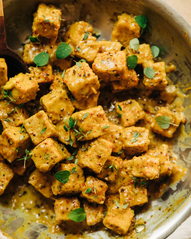 An up close, overhead shot of saucy chunks of tempeh in a skillet. The sauce is golden yellow and the tempeh is garnished with fresh thyme and oregano leaves.