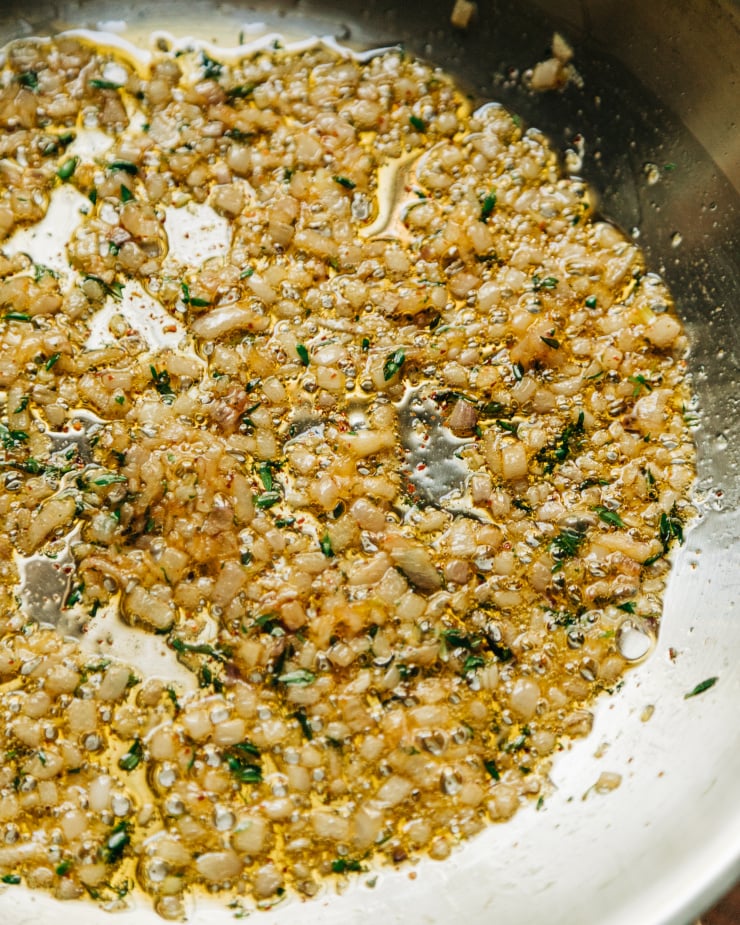 A 3/4 angle shot of sizzling diced shallots, thyme, and chillies in an oiled skillet.