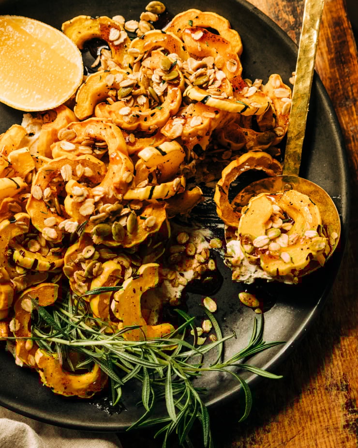 An overhead shot of roasted delicata squash pieces topped with rosemary granola and spiced maple syrup; all on top of a matte black plate with a wedge of lemon to the side.