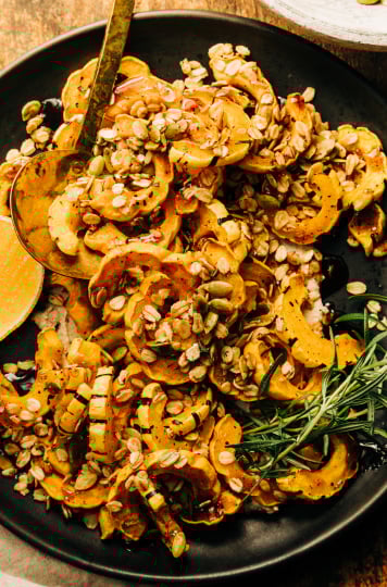 An overhead shot of roasted delicata squash pieces topped with rosemary granola and spiced maple syrup; all on top of a matte black plate with a wedge of lemon to the side.