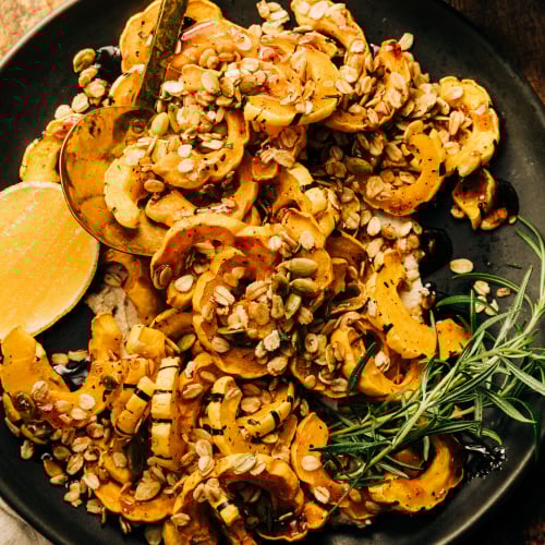 An overhead shot of roasted delicata squash pieces topped with rosemary granola and spiced maple syrup; all on top of a matte black plate with a wedge of lemon to the side.