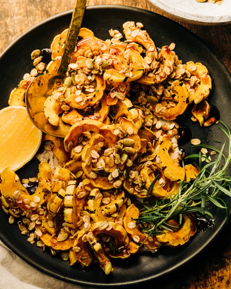 An overhead shot of roasted delicata squash pieces topped with rosemary granola and spiced maple syrup; all on top of a matte black plate with a wedge of lemon to the side.