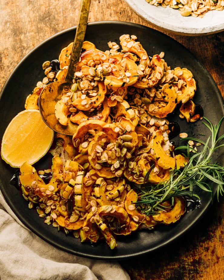 An overhead shot of roasted delicata squash pieces topped with rosemary granola and spiced maple syrup; all on top of a matte black plate with a wedge of lemon to the side.