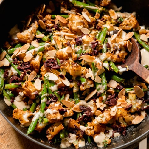 An up close, overhead shot of a sautéed cauliflower and green bean dish that is topped with a creamy sauce, chopped olives, and sliced almonds.