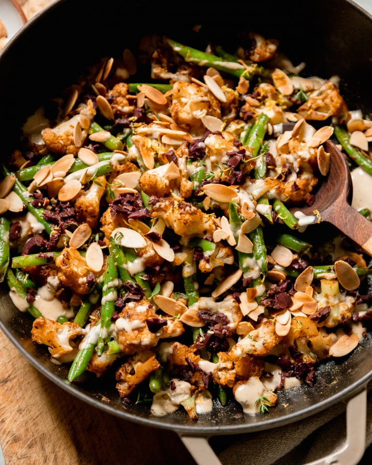 An up close, overhead shot of a sautéed cauliflower and green bean dish that is topped with a creamy sauce, chopped olives, and sliced almonds.