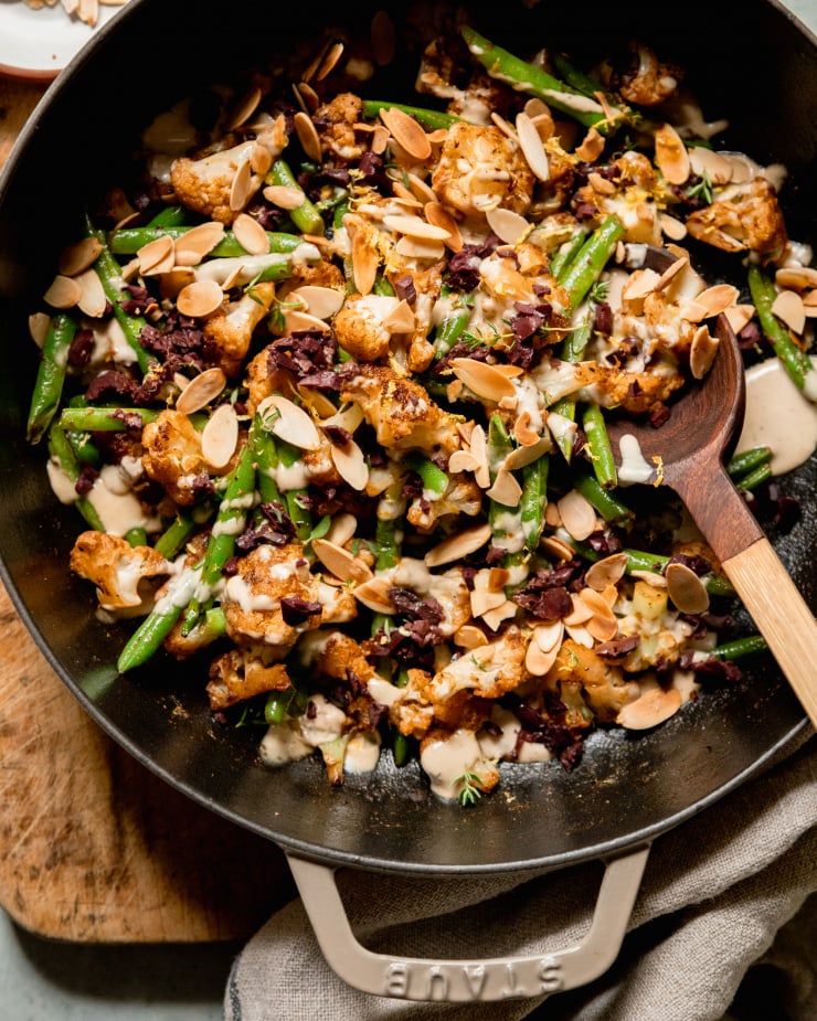 An up close, overhead shot of a sautéed cauliflower and green bean dish that is topped with a creamy sauce, chopped olives, and sliced almonds.