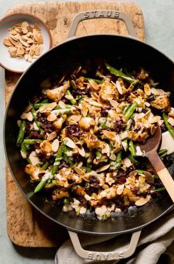 An overhead shot of a sautéed cauliflower and green bean dish that is topped with a creamy sauce, chopped olives, and sliced almonds. The dish is plated in a large braiser-style pot and is perched on top of a wood cutting board.
