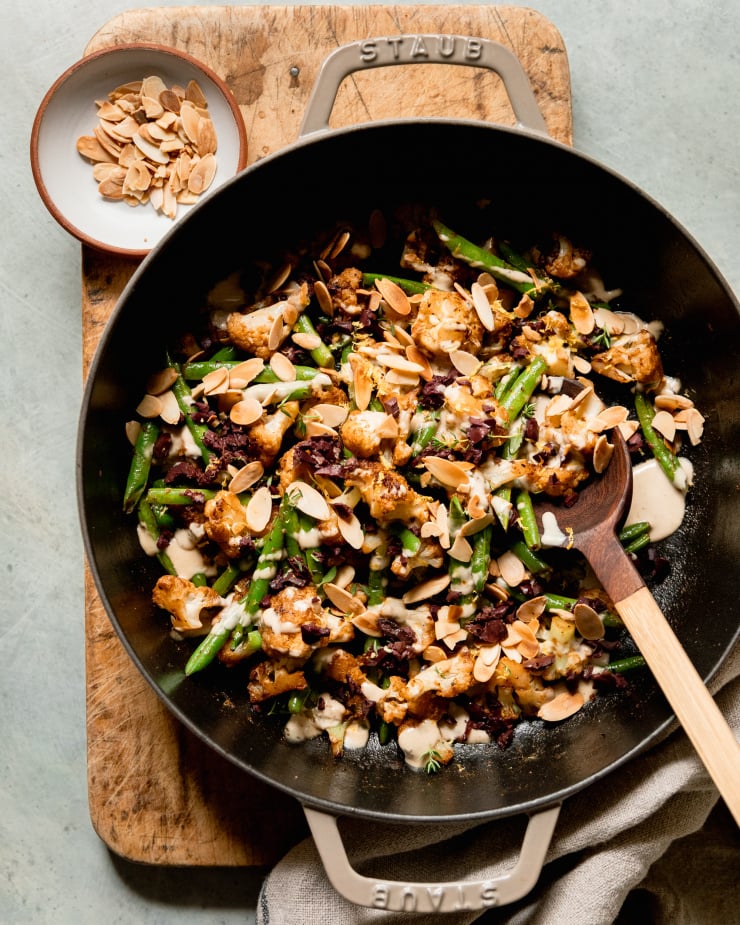 An overhead shot of a sautéed cauliflower and green bean dish that is topped with a creamy sauce, chopped olives, and sliced almonds. The dish is plated in a large braiser-style pot and is perched on top of a wood cutting board.