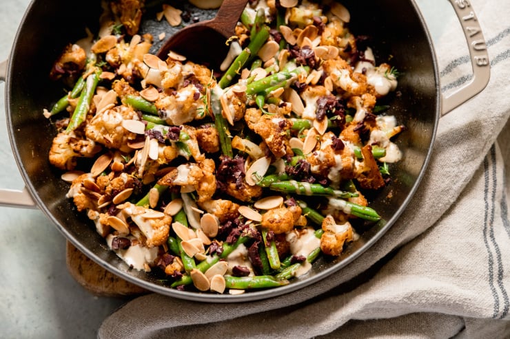 An up close, overhead shot of a sautéed cauliflower and green bean dish that is topped with a creamy sauce, chopped olives, and sliced almonds.