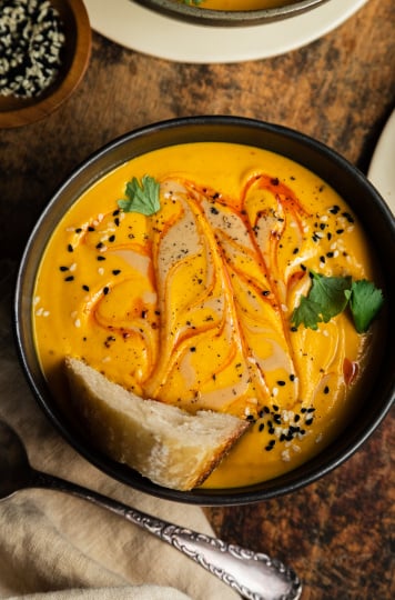 An overhead shot of a bowl of vibrant and creamy orange soup that is swirled with a red slick of oil on top. The soup has a crust of bread in it and is also garnished with sesame seeds and cilantro.