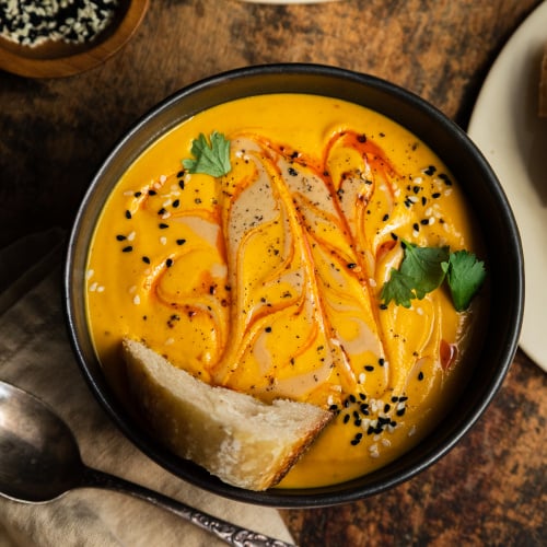 An overhead shot of a bowl of vibrant and creamy orange soup that is swirled with a red slick of oil on top. The soup has a crust of bread in it and is also garnished with sesame seeds and cilantro.