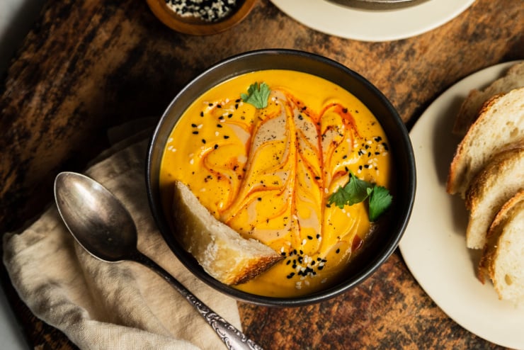 An overhead shot of a bowl of vibrant and creamy orange soup that is swirled with a red slick of oil on top. The soup has a crust of bread in it and is also garnished with sesame seeds and cilantro.