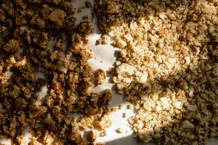 An overhead shot of cauliflower rice and bits of tofu, both coated in spices, on a parchment-lined baking sheet, pre-roasting.