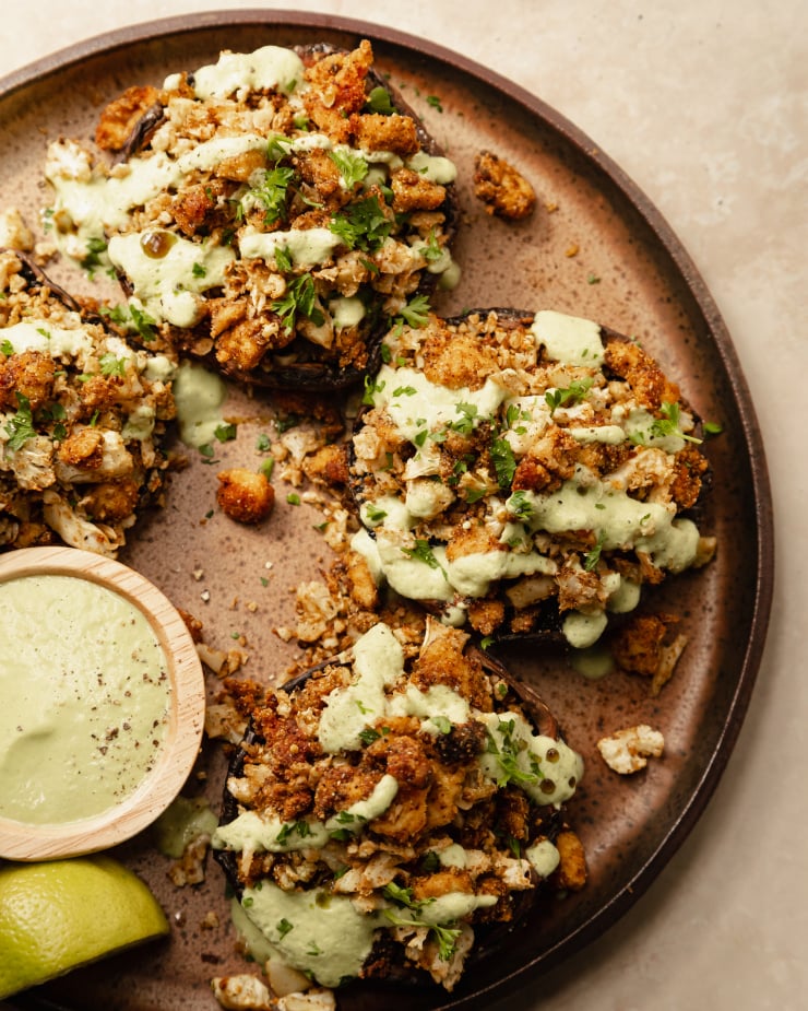 An overhead shot of some stuffed portobello mushrooms drizzled with a pale green sauce on top of a speckled brown plate. Part of a roundup of vegan tofu recipes.