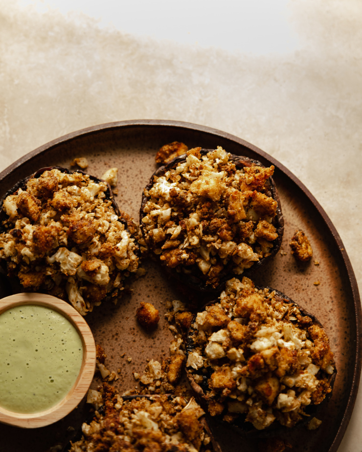 An overhead shot of some stuffed portobello mushrooms on a speckled brown plate. A small bowl of green sauce is seen to the side.