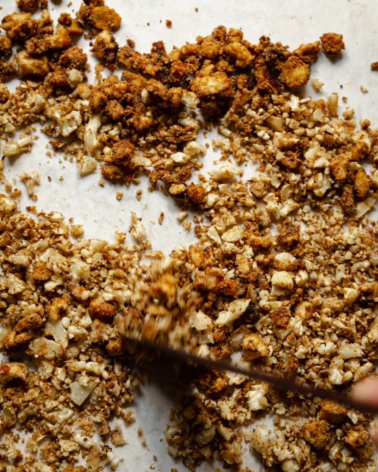 An overhead shot of roasted cauliflower rice and crispy coated bits of tofu being stirred up on a parchment-lined baking sheet.