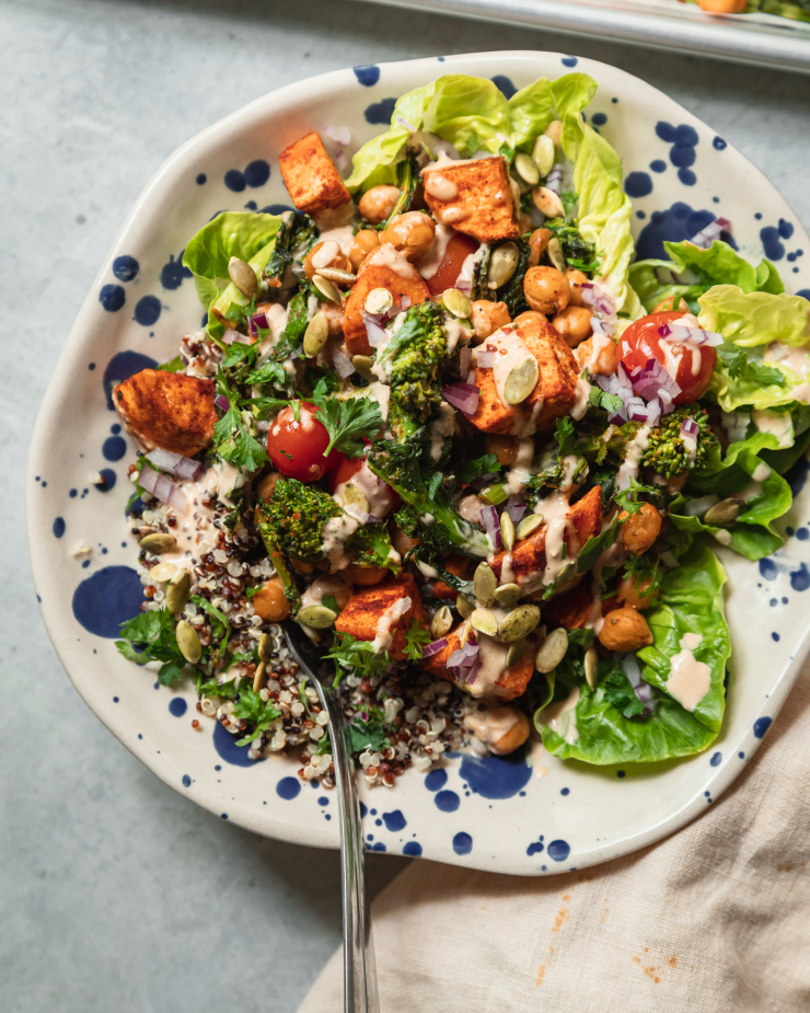 An overhead shot of a quinoa grain bowl in a blue splattered bowl. The quinoa is accompanied by roasted sweet potatoes, broccoli rabe, cherry tomatoes, chickpeas, greens, and drizzles of peanut sauce.