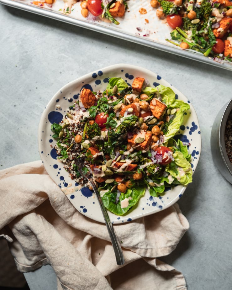 An overhead shot of a quinoa grain bowl in a blue splattered bowl. The quinoa is accompanied by roasted sweet potatoes, broccoli rabe, cherry tomatoes, chickpeas, greens, and drizzles of peanut sauce.