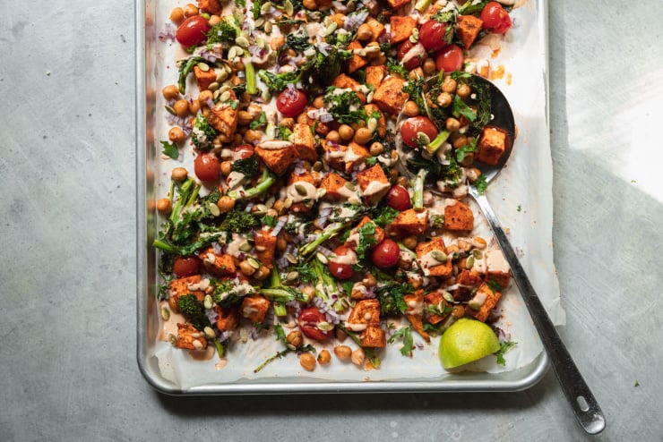 An overhead shot of roasted sweet potatoes, broccoli rabe, chickpeas, and cherry tomatoes drizzled with a creamy peanut sauce. There are garnishes of diced red onion and pumpkin seeds on top as well. All components are contained in a sheet pan.