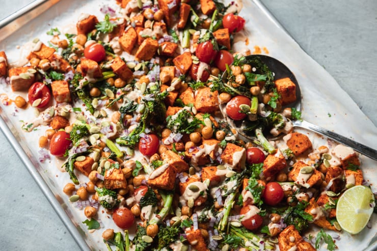 An up close, overhead shot of roasted sweet potatoes, broccoli rabe, chickpeas, and cherry tomatoes drizzled with a creamy peanut sauce. There are garnishes of diced red onion and pumpkin seeds on top as well. All components are contained in a sheet pan.