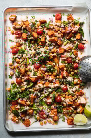An overhead shot of roasted sweet potatoes, broccoli rabe, chickpeas, and cherry tomatoes drizzled with a creamy peanut sauce. There are garnishes of diced red onion and pumpkin seeds on top as well. All components are contained in a sheet pan.