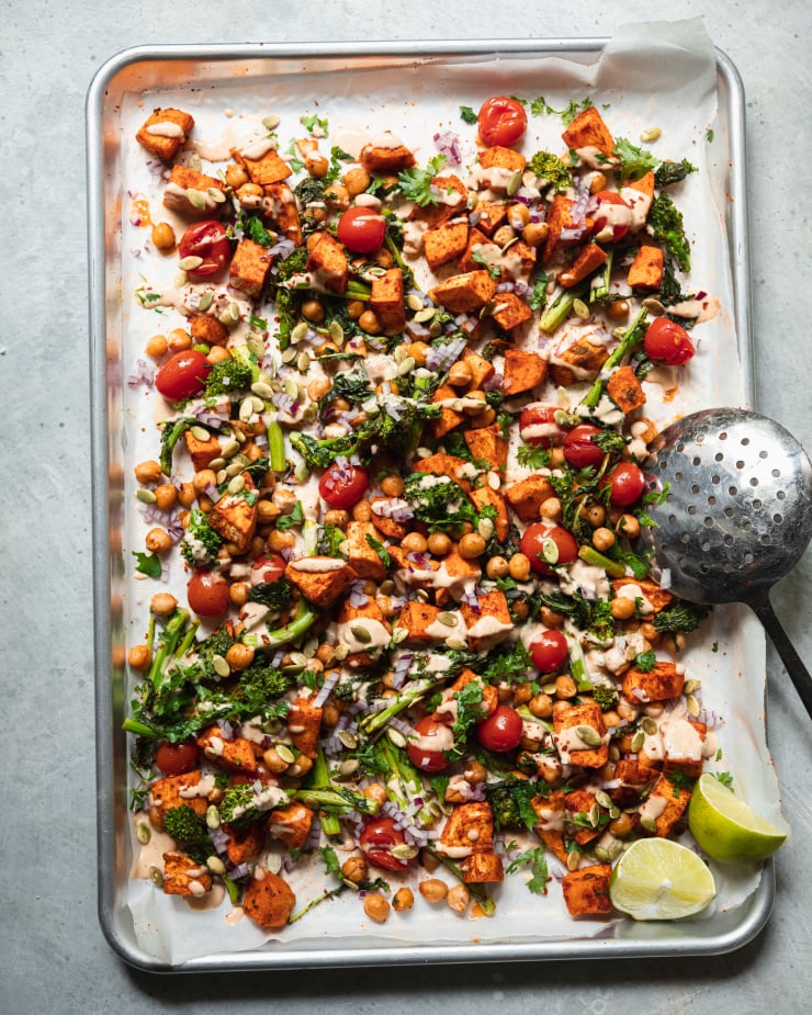 An overhead shot of roasted sweet potatoes, broccoli rabe, chickpeas, and cherry tomatoes drizzled with a creamy peanut sauce. There are garnishes of diced red onion and pumpkin seeds on top as well. All components are contained in a sheet pan.