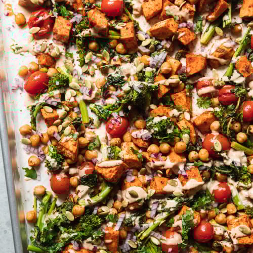 An up close, overhead shot of roasted sweet potatoes, broccoli rabe, chickpeas, and cherry tomatoes drizzled with a creamy peanut sauce. There are garnishes of diced red onion and pumpkin seeds on top as well. All components are contained in a sheet pan.