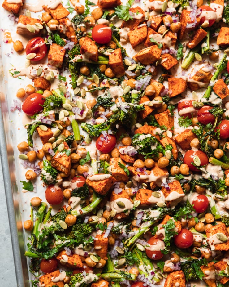 An up close, overhead shot of roasted sweet potatoes, broccoli rabe, chickpeas, and cherry tomatoes drizzled with a creamy peanut sauce. There are garnishes of diced red onion and pumpkin seeds on top as well. All components are contained in a sheet pan.