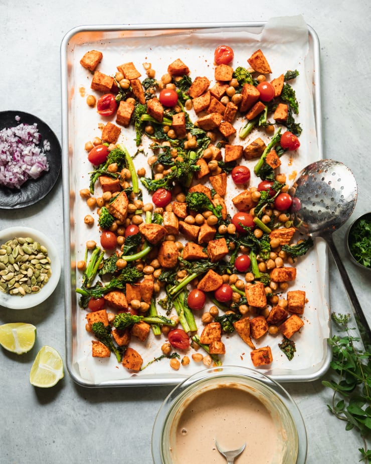 An overhead shot of roasted sweet potatoes, broccoli rabe, cherry tomatoes, and chickpeas on a sheet pan with some garnishes in small bowls nearby.