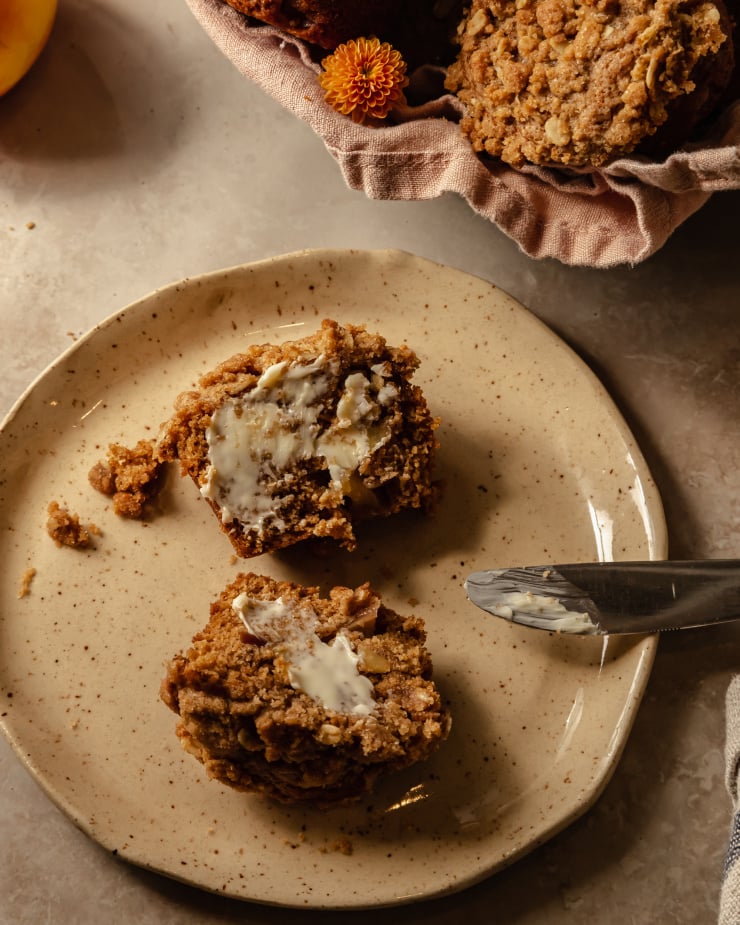 Image shows a vegan apple crisp muffin split in half and spread with swoops of vegan butter on both sides.