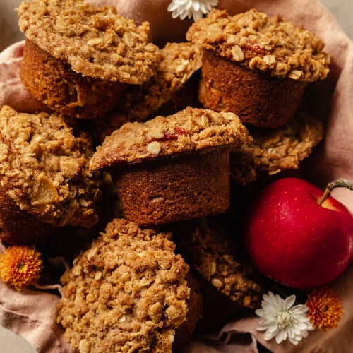 An overhead shot of crumble topped muffins set inside of a pink linen in a bowl. An apple is shown to the side.