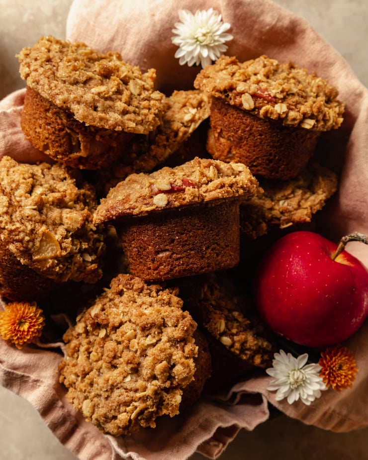 An overhead shot of crumble topped muffins set inside of a pink linen in a bowl. An apple is shown to the side.