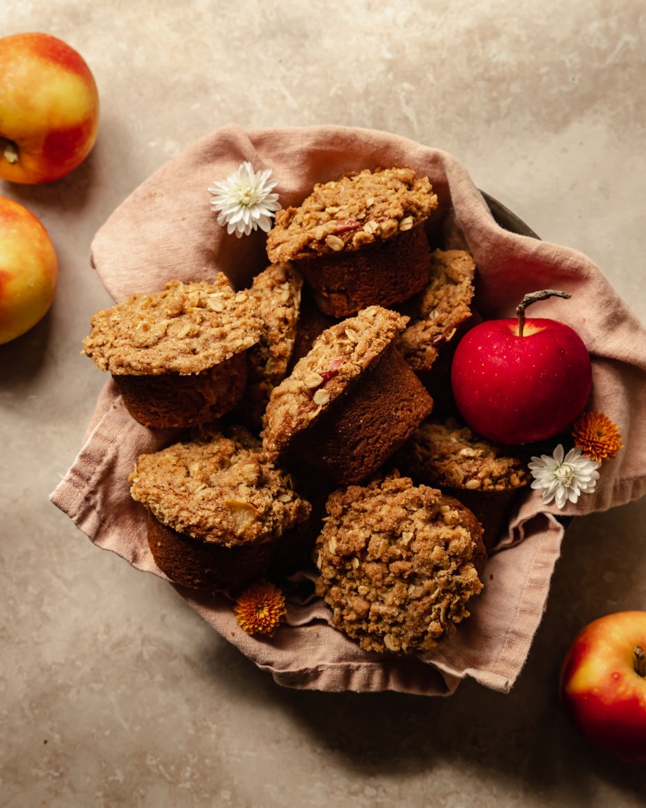 An overhead shot of crumble topped muffins set inside of a pink linen in a bowl. An apple is shown to the side.
