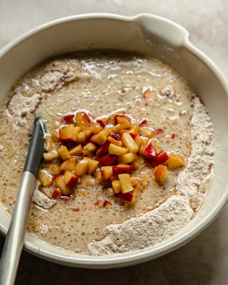 A 3/4 angle image shows cooked diced apples being added to muffin batter in a large white bowl.