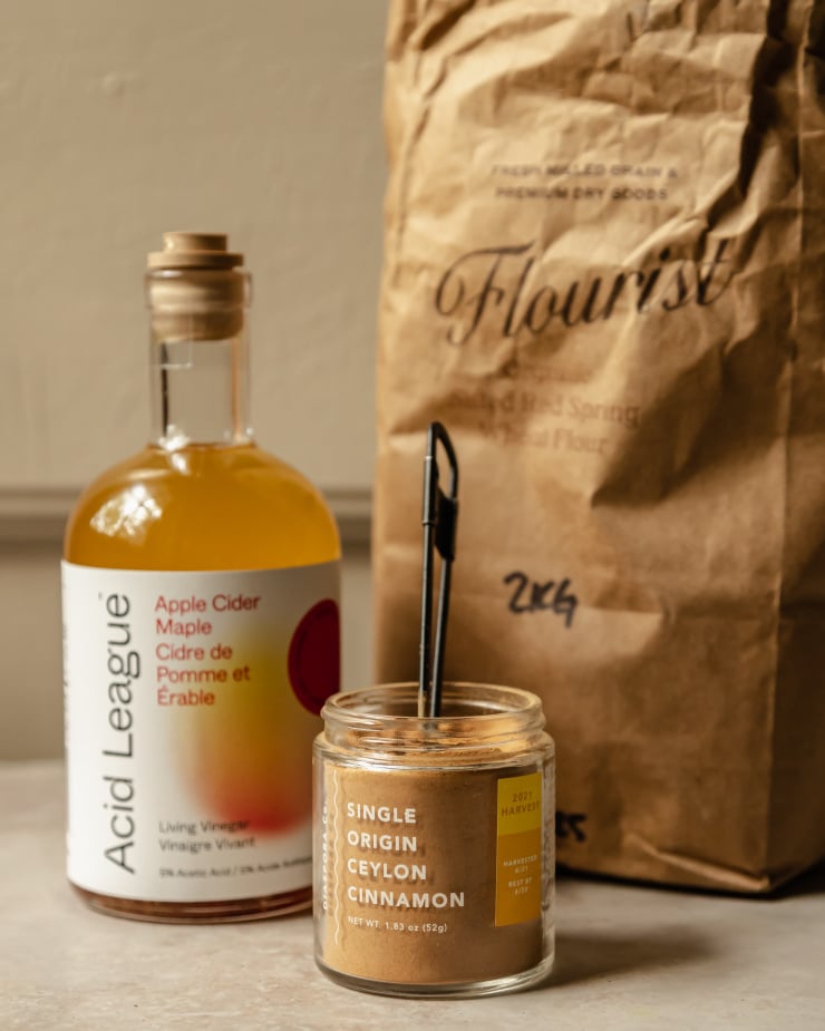 A head-on shot of a jar of ground cinnamon, a bottle of apple cider vinegar and a brown paper bag of flour.