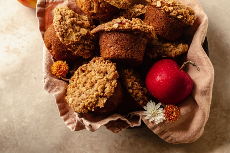 An overhead shot of crumble topped muffins set inside of a pink linen in a bowl. An apple is shown to the side.