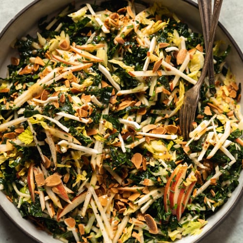 An up close, overhead shot of an apple kale salad with toasted almonds, photographed in a white ceramic dish that is rimmed with matte black.