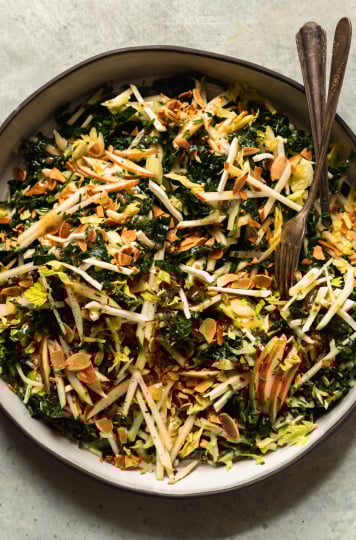 An overhead shot of an apple kale salad with toasted almonds, photographed in a white ceramic dish that is rimmed with matte black.