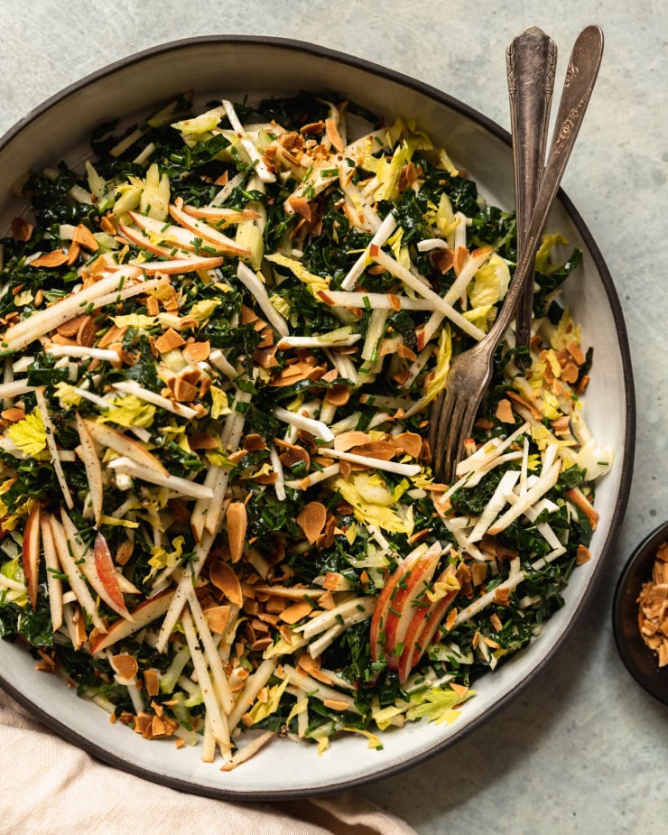 An overhead shot of an apple kale salad with toasted almonds, photographed in a white ceramic dish that is rimmed with matte black.