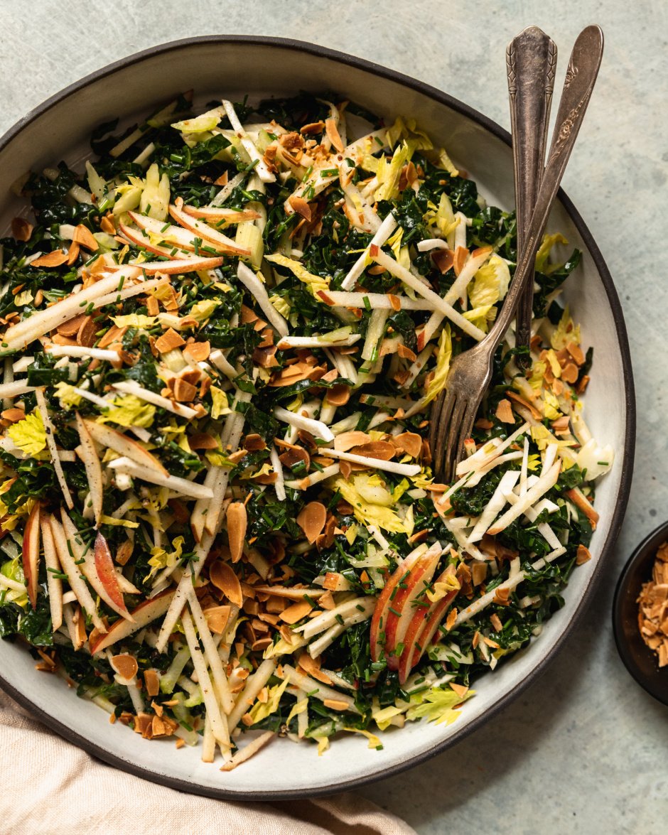 An overhead shot of an apple kale salad with toasted almonds, photographed in a white ceramic dish that is rimmed with matte black.
