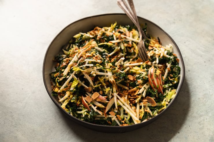 An 3/4 shot of an apple kale salad with toasted almonds, photographed in a white ceramic dish that is rimmed with matte black.